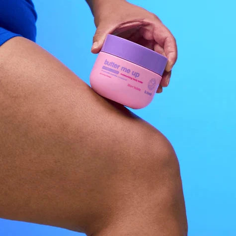 Butter Me Up hydraterende body butter pot in hand, met soepel glanzend been op blauwe achtergrond.